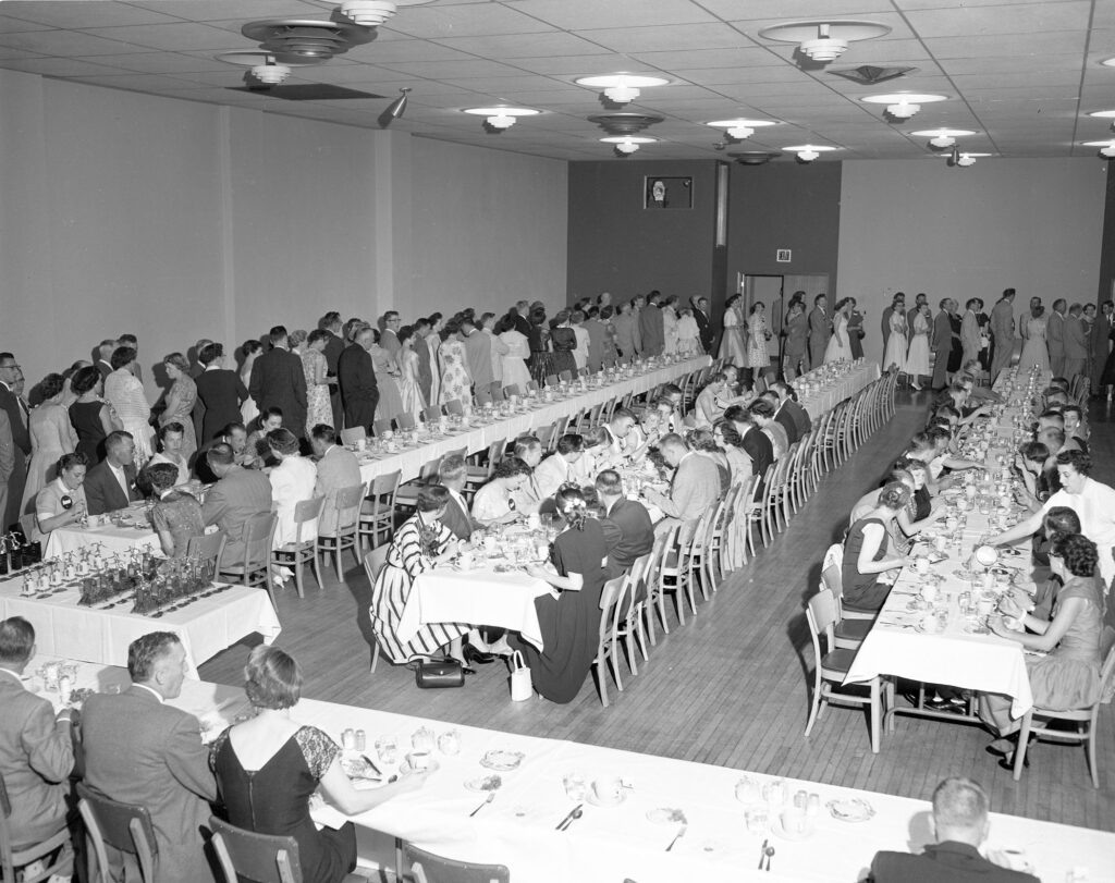 A black and white photograph shows people form in a line next to rows of long tables as others eat and drink at the function.