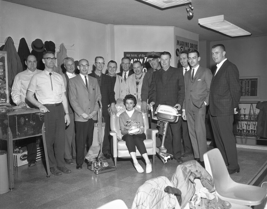 A black and white photograph shows a group of men standing around a woman in a chair.