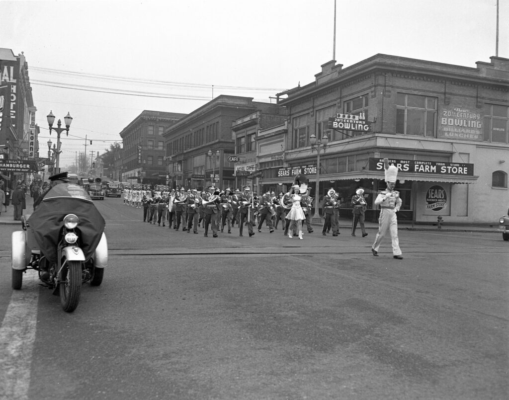 A black and white photograph shows a marching band pass a bowling alley during a parade.