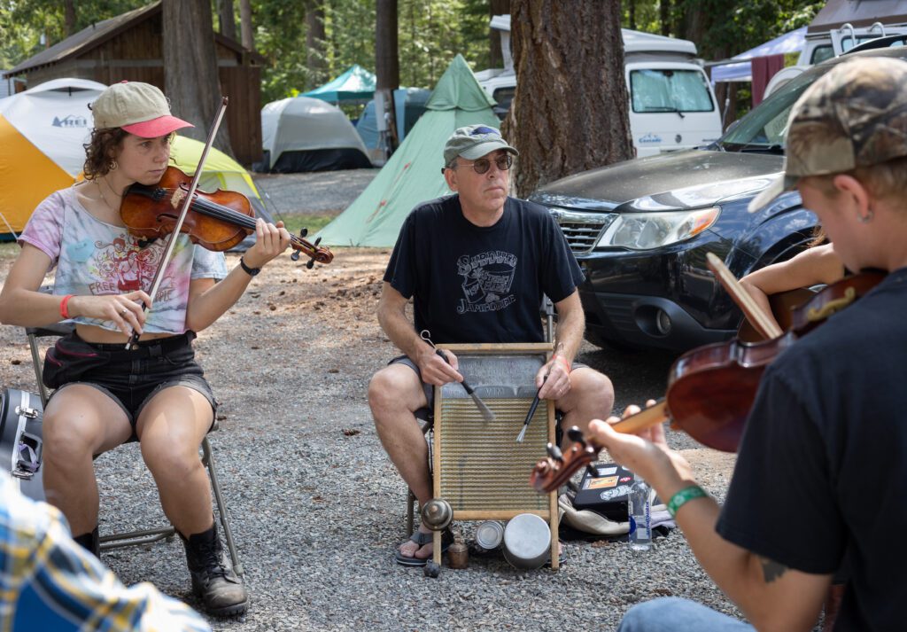 Attendees play instruments in a circle next to tents and their cars.