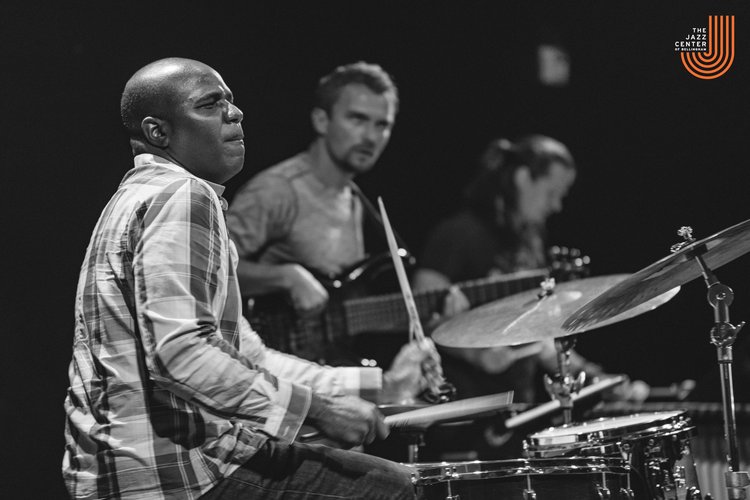 A black and white photo of D’Vonne Lewis plays the drums next to bandmates.