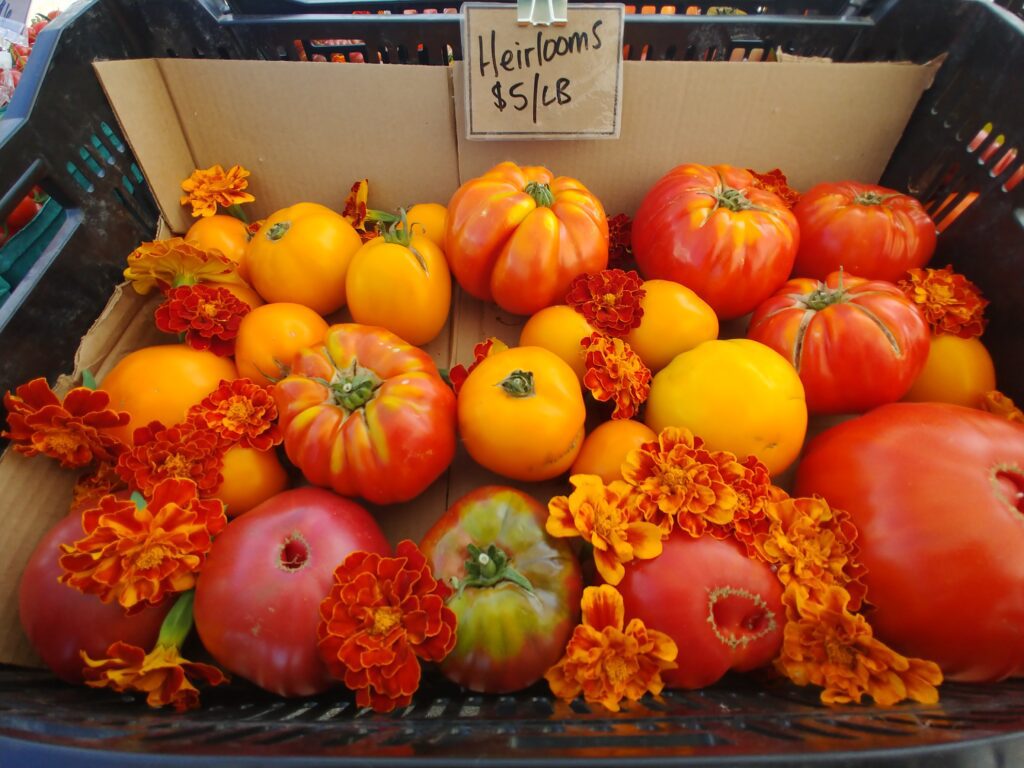 Heirloom tomatoes in a crate with flowers and a sign that shows h ow much they cost per pound.