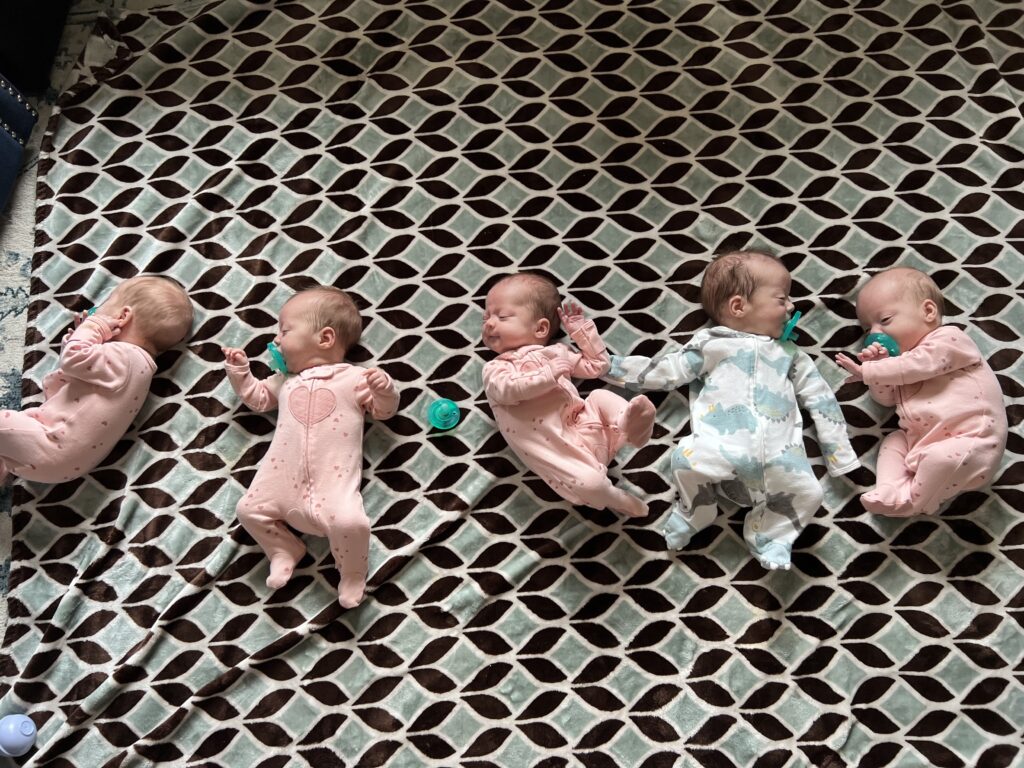 Five babies, quintuplets, lay on the floor next to their siblings.