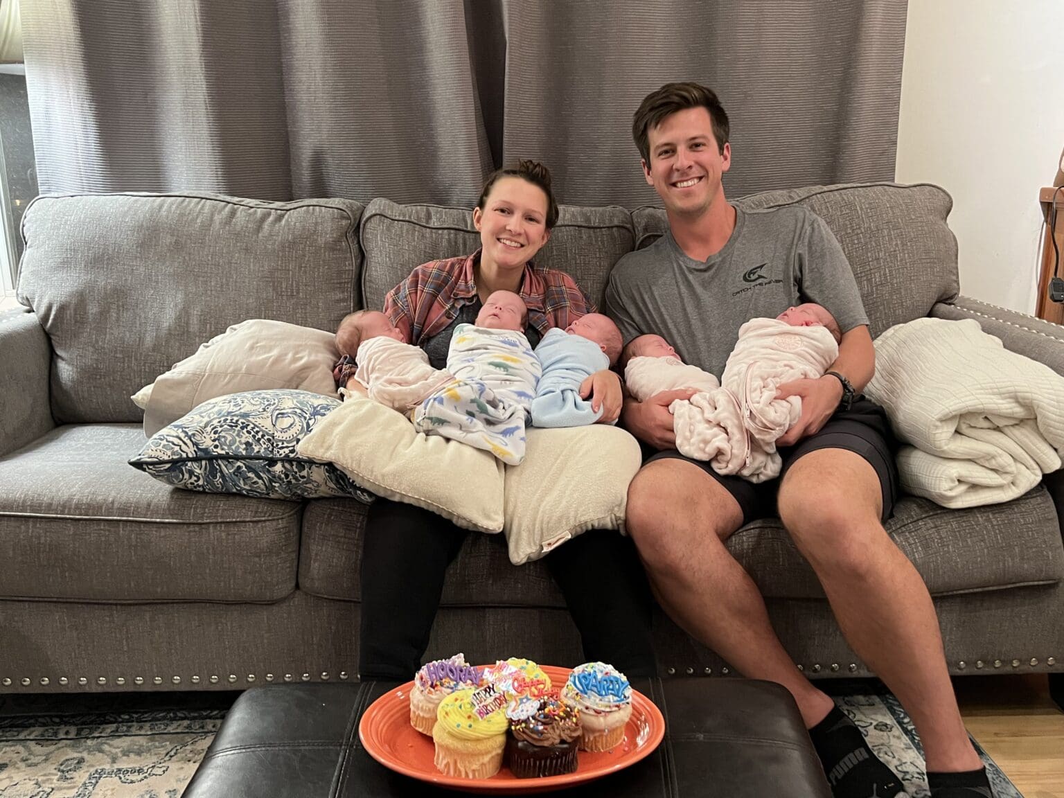 Stephanie and Graham Freels sitting on a couch in their living room holds up their quintuplets as they smile for the camera.