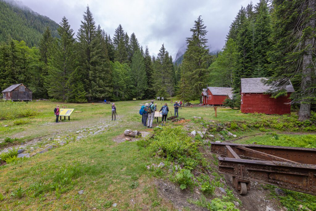 Hikers roam through the Monte Cristo mining site that has an informative board in the center surrounded by small red buildings.