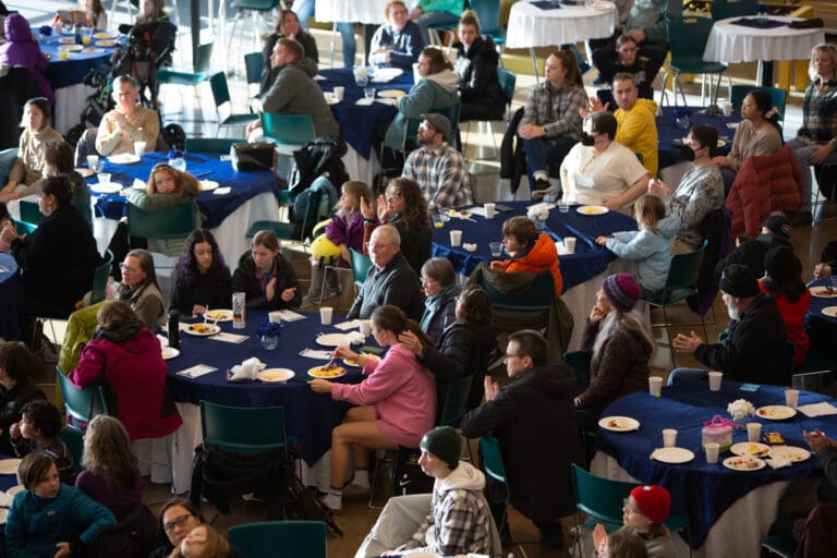 A crowd of people sitting at several tables covered in dark blue cloths cheering and watching.