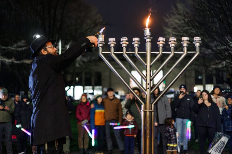 Rabbi Avremi Yarmush lighting a menorah as a crowd of spectators watch.