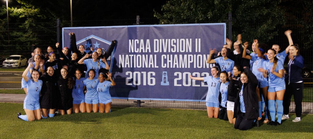 Western women's soccer team poses next to the banner of NCAA division National Champion.