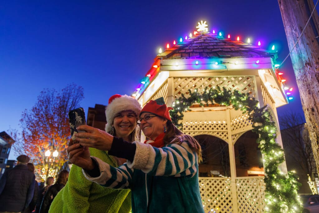 Lori Palmer-Thompson, dressed as Grinch, and Liz Wadsworth, dressed as an elf, take a selfie together with the decorated and lit up gazebo behind them.