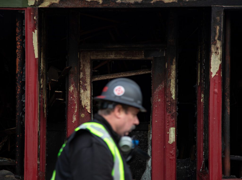 An official walks in front of the doorway to the burned Harris Avenue Cafe wearing a grey helmet, safety vest and a gas mask hanging below his chin.