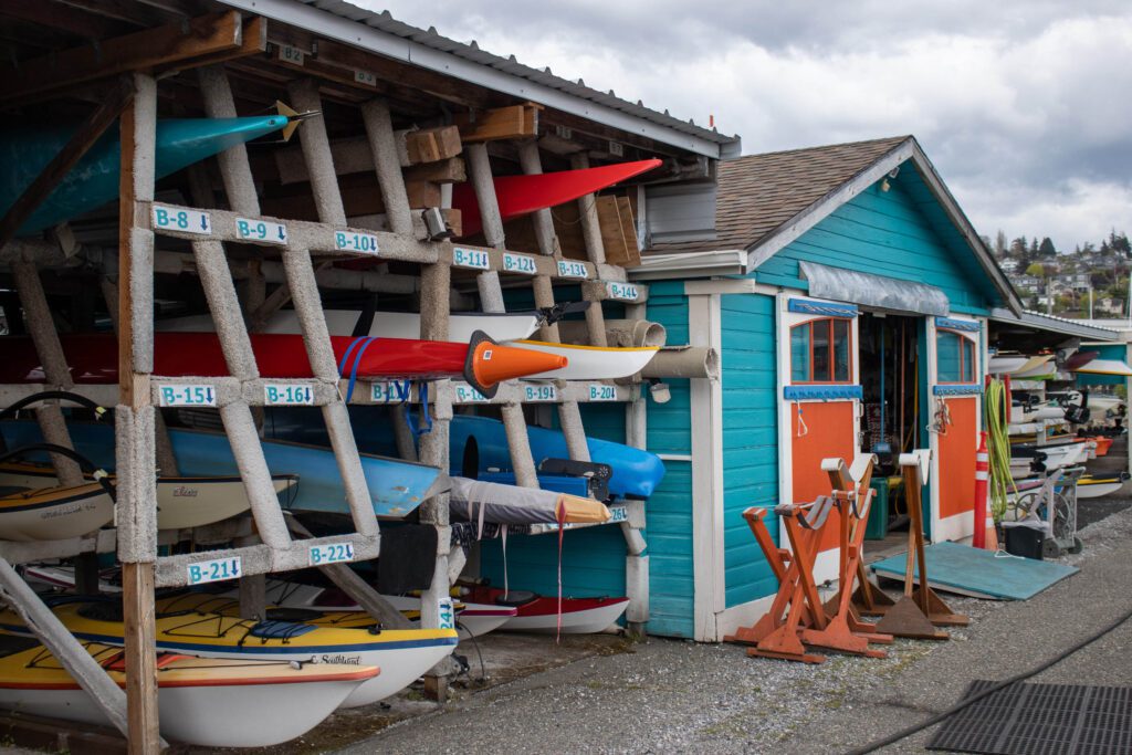 The colorful front of Community Boating Center has rows of paddleboards stacked on the side of the blue building.