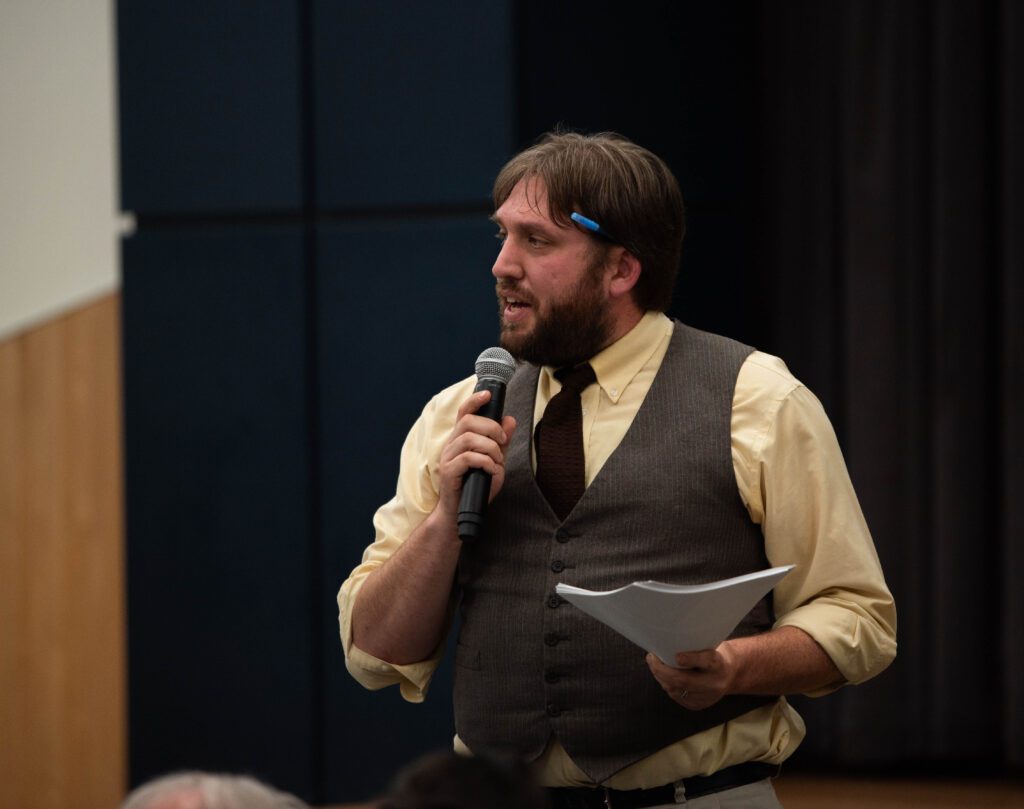 ABC Recycling Community Relations Manager Riley Sweeney talks through a microphone with a pen behind his ear as he holds a stack of papers with his free hand.