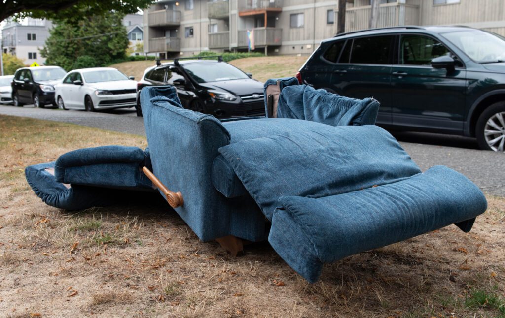 A tattered recliner in full repose sits on the side of 21st Street with parts of it lying on the ground next to it.