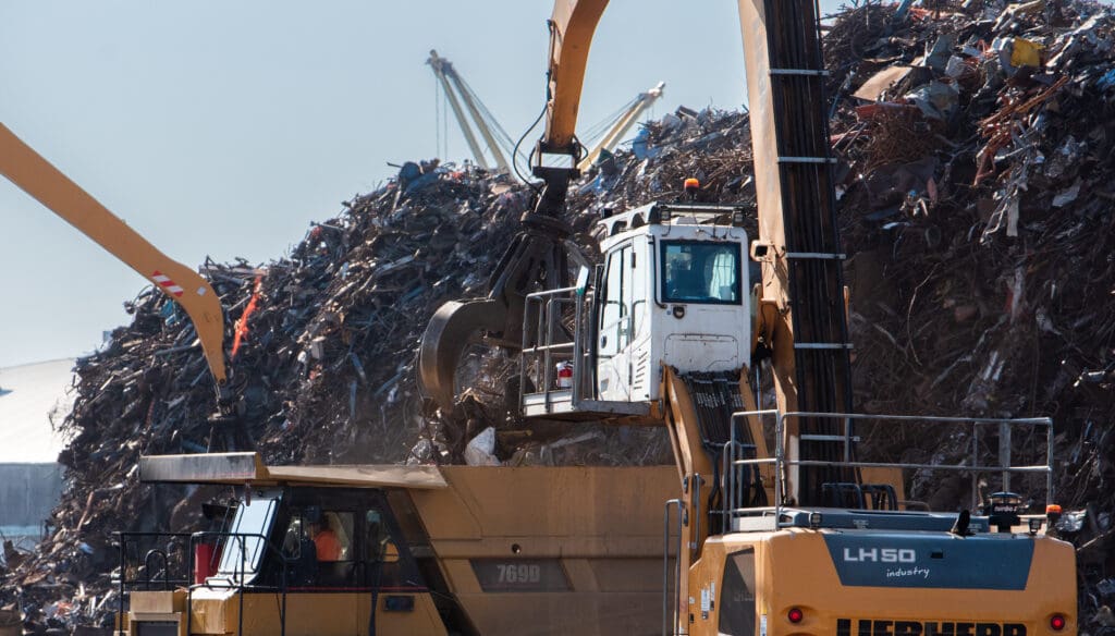 An excavator places scrap metal in a dump truck surrounded by other scrap piles.