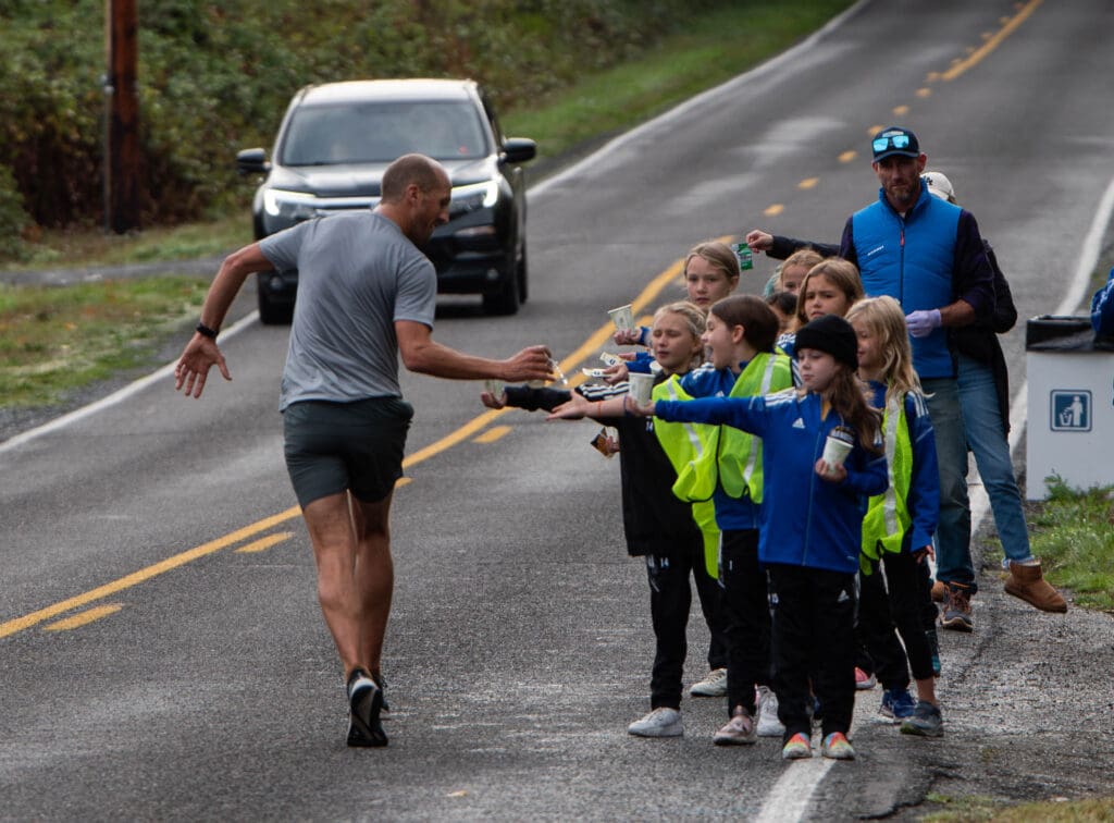 Scott Lipp receives water from a small huddle of members of the Whatcom FC Rangers who are reaching out for runners to grab the water from them as a car drives down from the other side of the road.