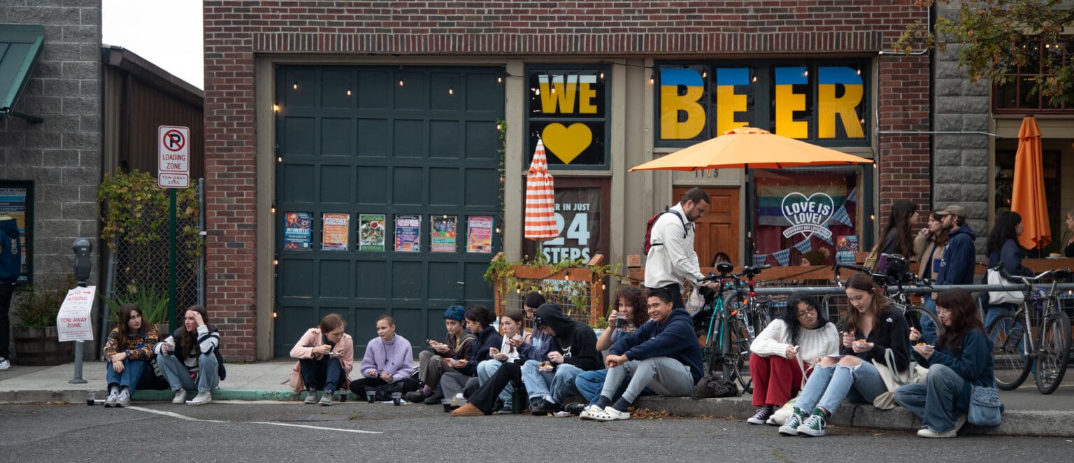 Groups of students eat free Boundary Bay Brewery macaroni cheese and sip on root beer on the curb outside the restaurant during the procession.