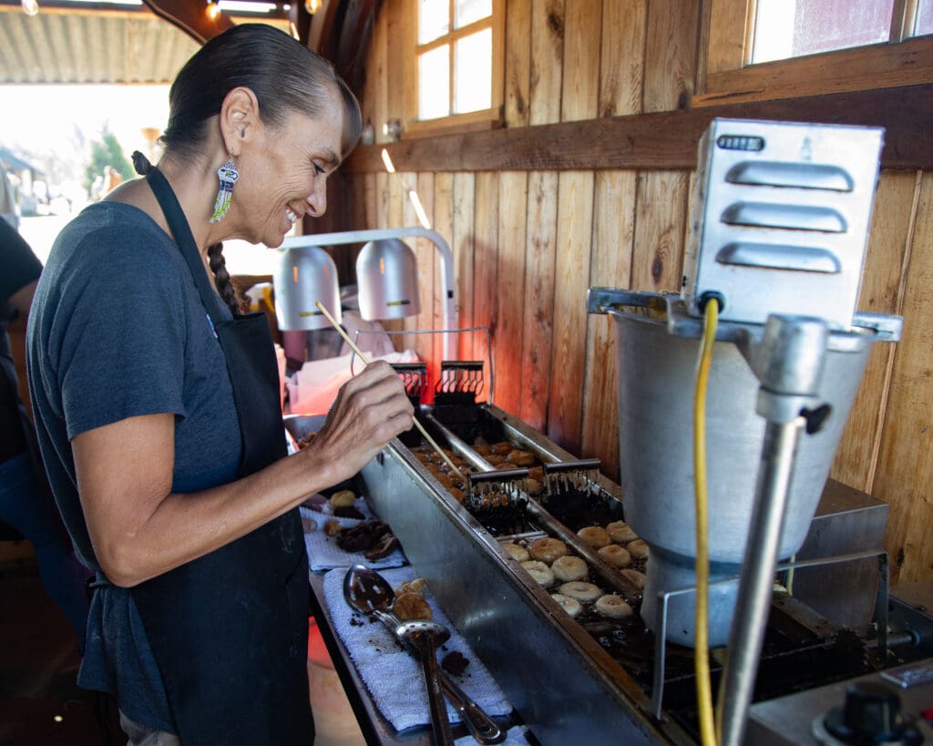 Tami Jefferson fries apple cider doughnuts with a smile while surrounded by metal tools that she uses to flip the doughnuts.