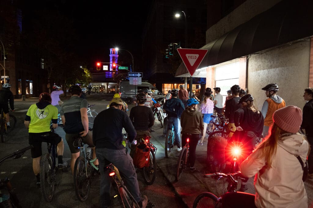 Bikers regrouping in a crowded huddle in the middle of the night as one biker has a shiny red light attached to their bike.