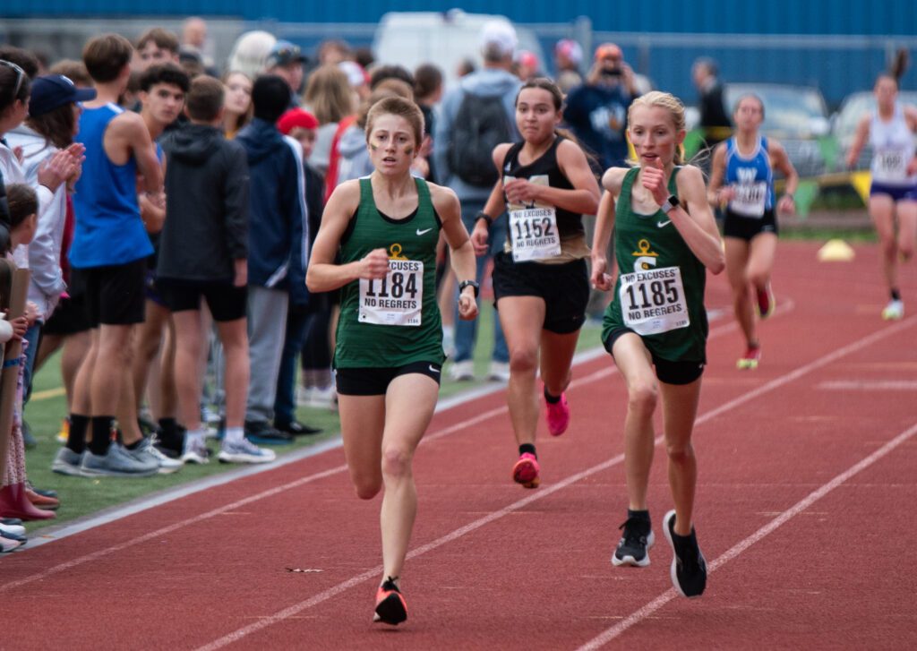 Sehome junior Abby Rusk, left, and freshman Lil Desler lead ahead of other runners on the red track field next to onlookers cheering for the participants.