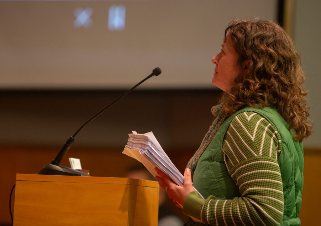 Tsena Paulson holds a stack of petitions signed by local residents behind the wooden podium where a microphone is placed.