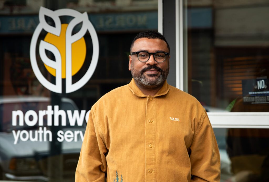 Northwest Youth Services Executive Director Jason McGill stands in front of the organization's offices wearing a bright yellow shirt to match the logo's color.