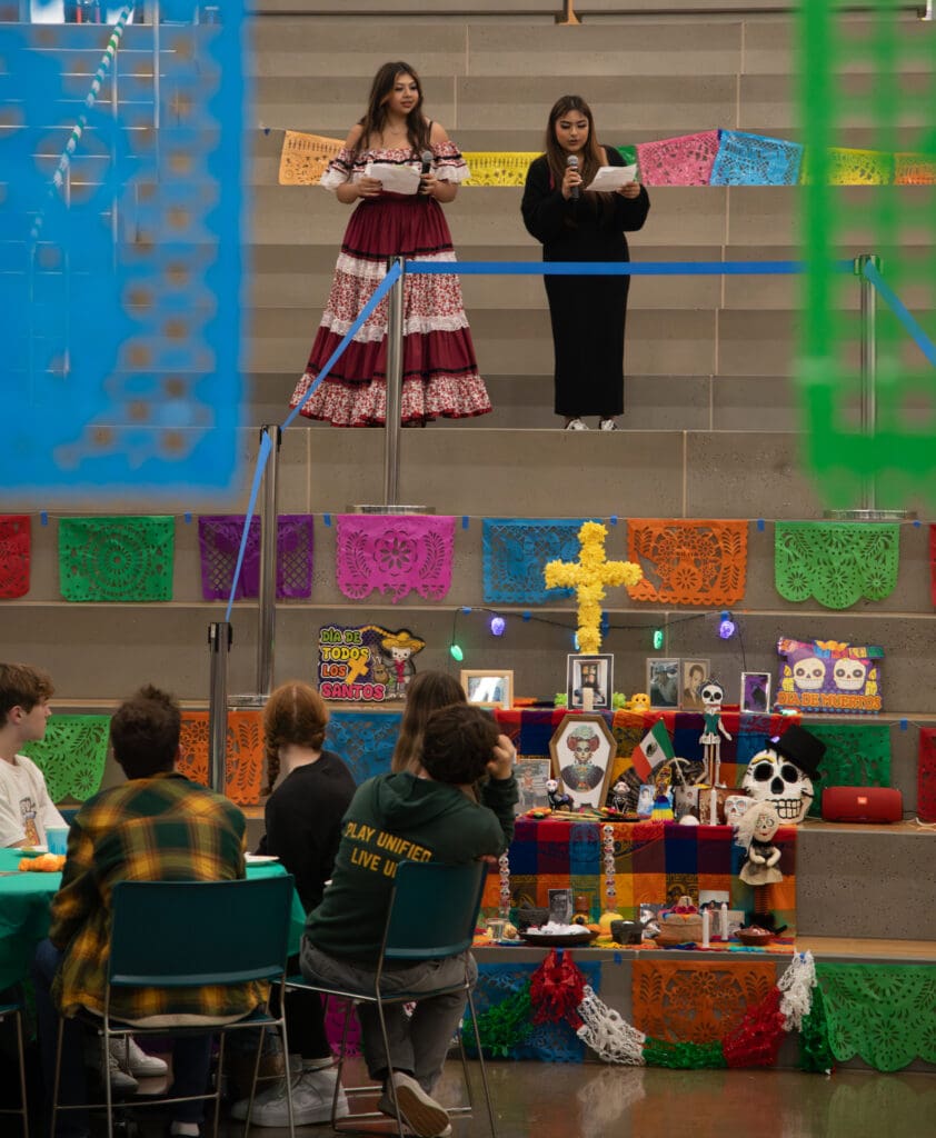 Seniors from the Latinx Club Analeigha Garza and Nayeli Arreola welcome attendees from above as a crowd sitting by them listens.