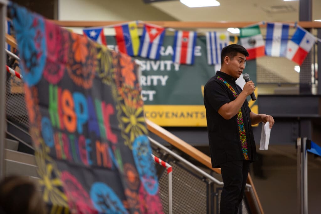 Squalicum High School Spanish teacher Hugo Santiago addresses the crowd next to a colorful banner with a mic in hand.