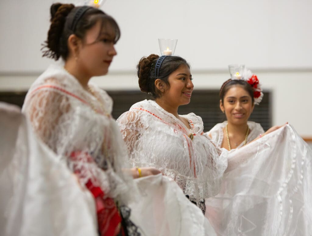 Students perform the traditional dance "La Bruja" while dressed in white dresses and keep balance for the glass candle holders on top of their heads.