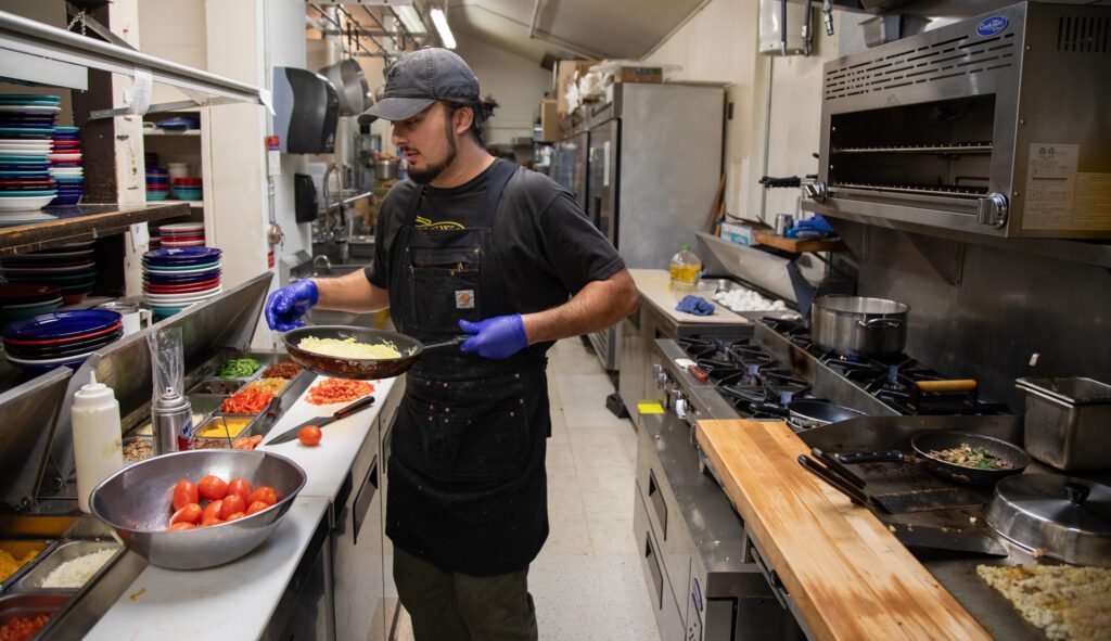 Dylan Swann cooks a Mediterranean omelet in the kitchen as he holds the pan next to trays full of fresh ingredients.