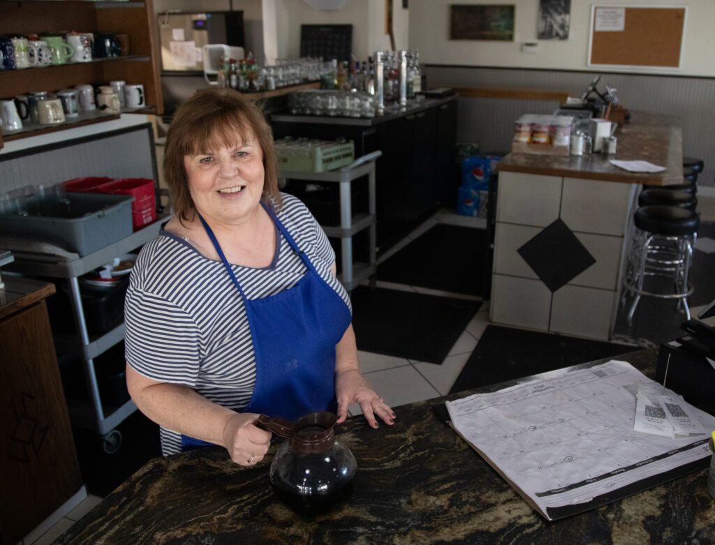 Sheryl Johnson, wearing a blue apron, stands with a carafe of hot coffee behind the bar.