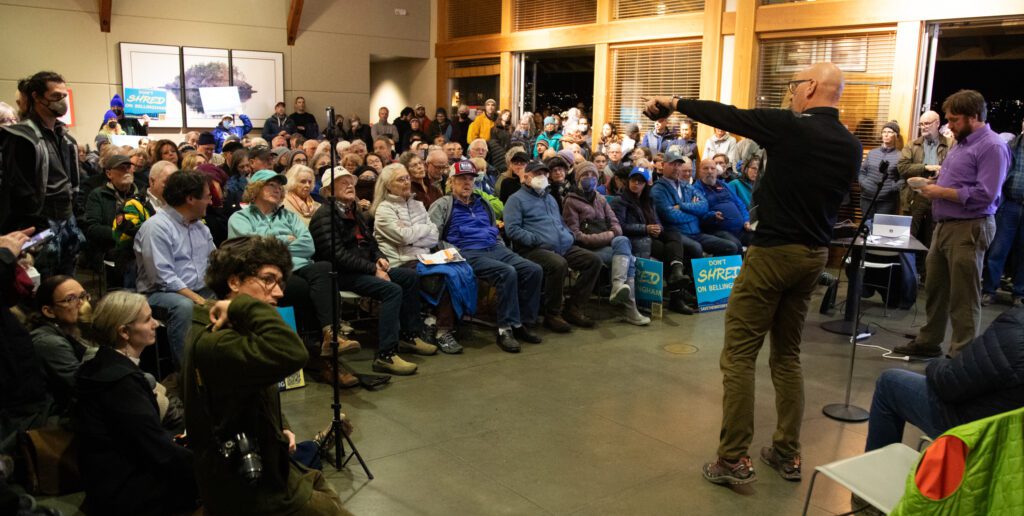 Approximately 300 people sit and stand in front of the speaker, some holding signs.