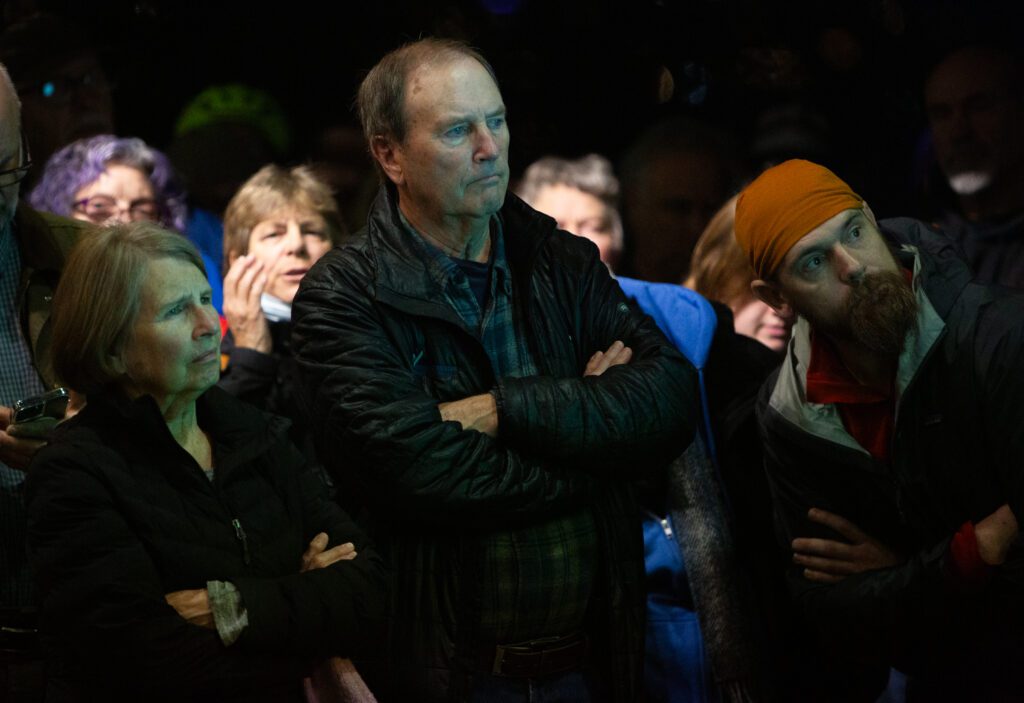 Three attendees surrounded by others are listening with their arms crossed.