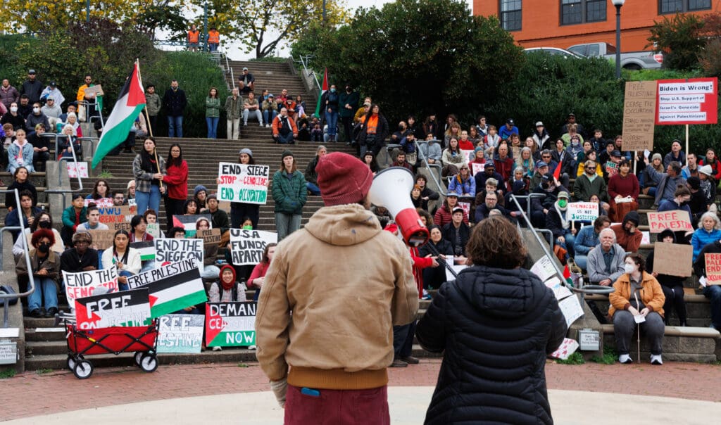 A view of the backs of the speakers as they address the crowd sitting on the steps with different signs and flags in support.