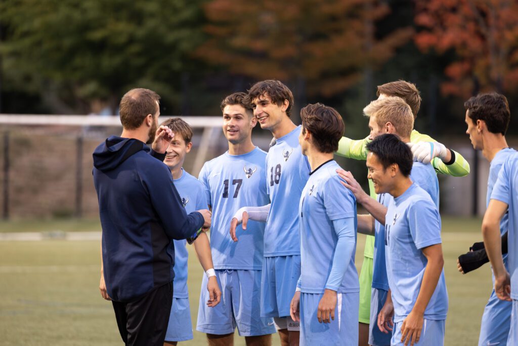 Western Washington University men's soccer players huddle around assistant coach Chuck Pitts with wide smiles.