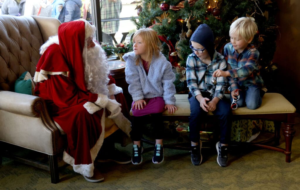 Santa talks with Olivia Long, 4, Logan Long, 6, and Oliver Stolle, 6 who are sitting on a yellow seat in front of the christmas tree.