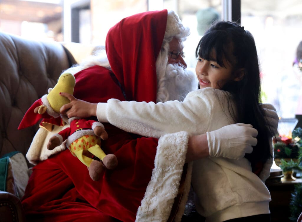 Ayiani Rubi gives Santa a hug with a yellow stuffed toy in hand.