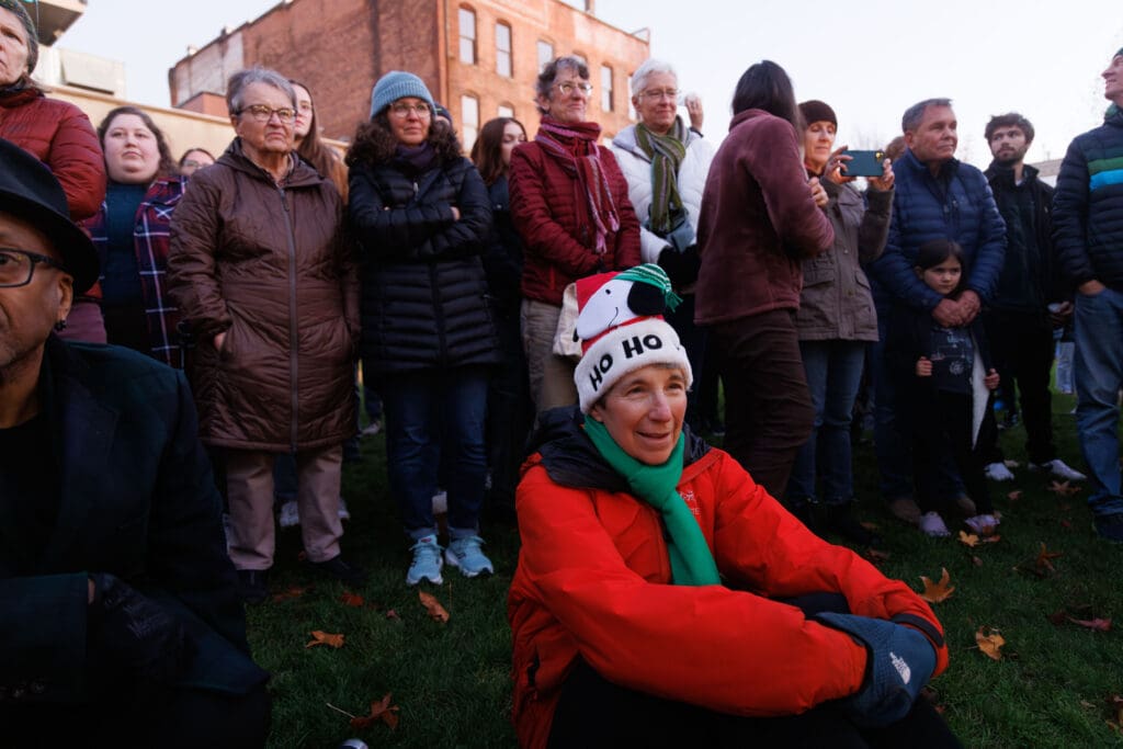 Leslee Probasco listens to the music of the Bellingham High Showstoppers choir by sitting on the grass wearing a Snoopy santa hat as other spectators stand behind her and watch.