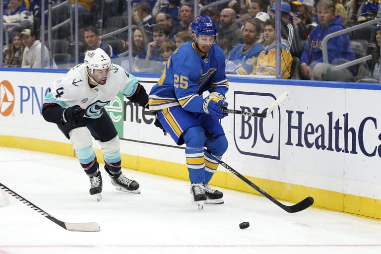 Seattle Kraken's Justin Schultz (4) and St. Louis Blues' Jordan Kyrou (25) battle for the puck next to the bleachers full of spectators.