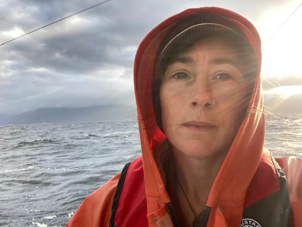 Author and commercial fisherman Tele Aadsen out at sea wearing a red hoodie and a beanie as the sun shines from behind her.