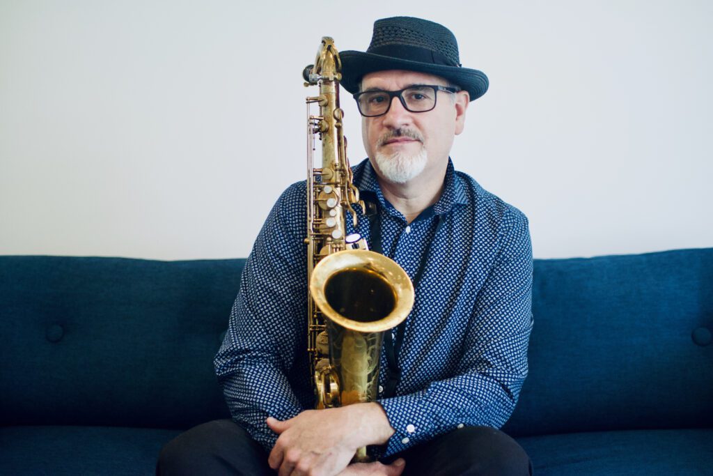 Vancouver jazz saxophonist Mike Allen poses for a photo with his saxophone while sitting on a blue couch.