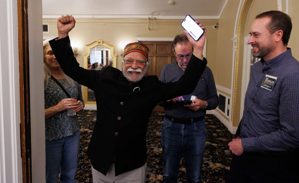 Whatcom County Executive incumbent Satpal Sidhu reacts next to county council candidate Jon Scanlon with his hands in the air, one of which is holding onto a phone with the results.