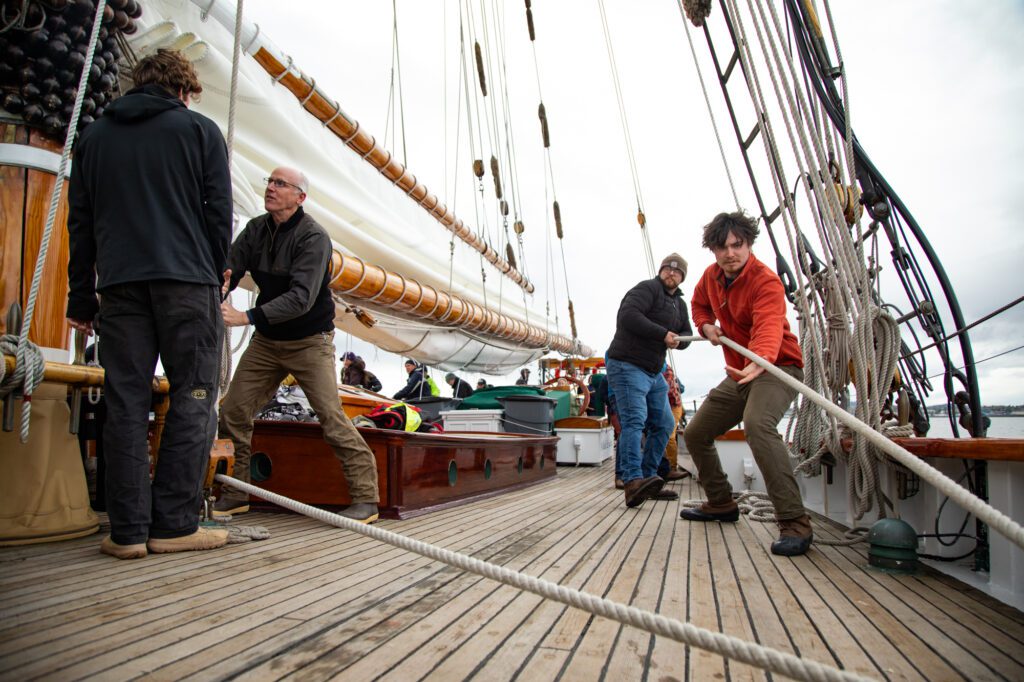 The main sail begins to rise as crew members pull the lines through a pulley system.