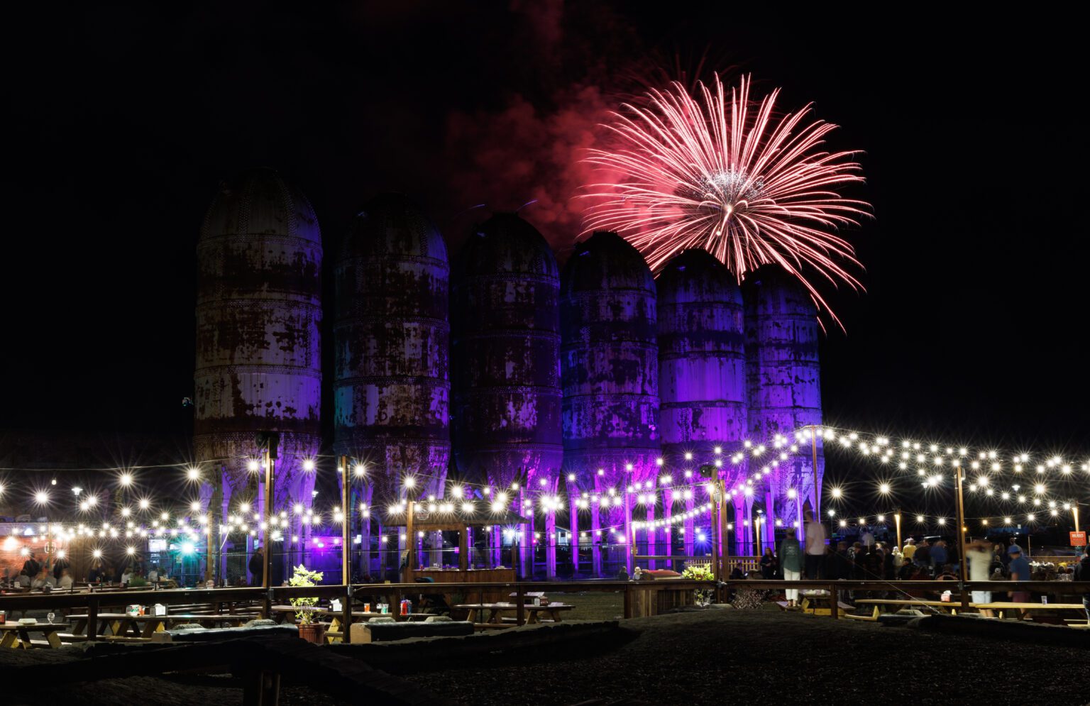 Fireworks explode behind the old containers as people watch from the outdoor seating of a restaurant.