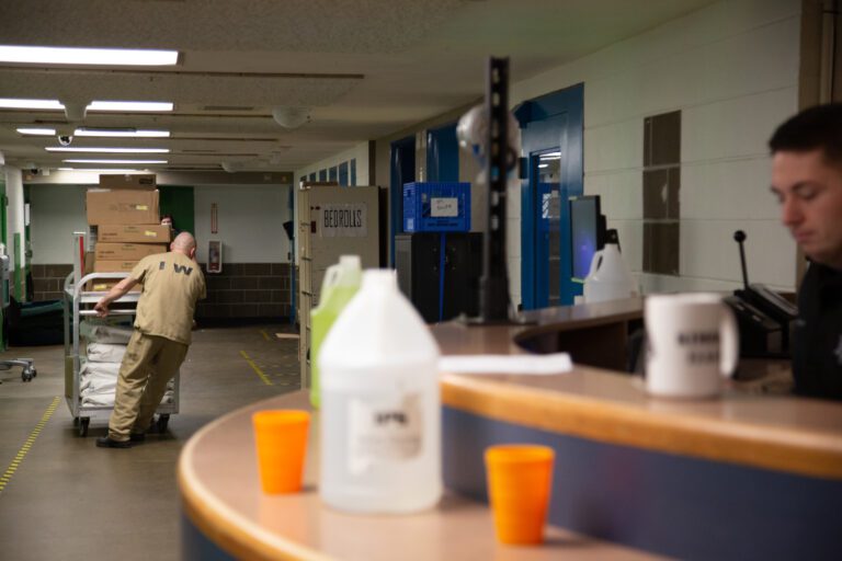 An inmate pulls a trolley of items as a guard sits watching from a reception desk.