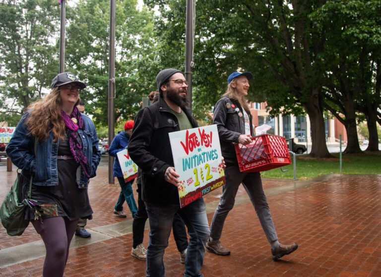 Members of Community First Whatcom walk with signs and a red crate filled with petitions for people to sign.