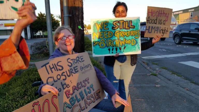 Two people, one sitting, hold signs in front of an intersection.