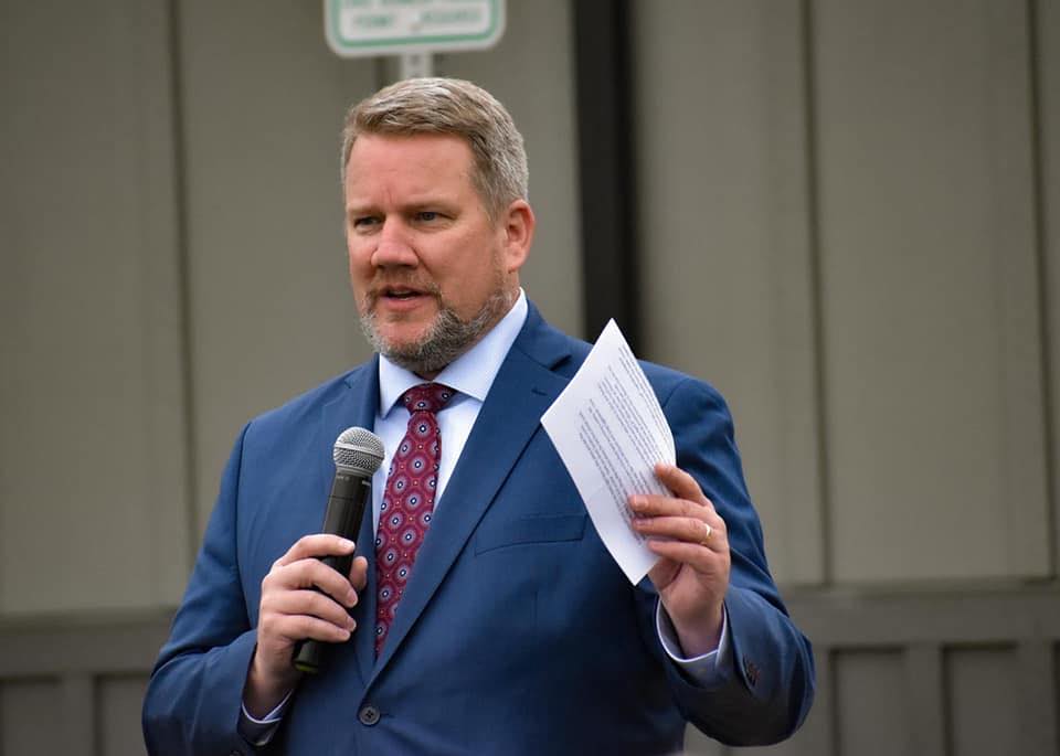 A bearded man wears a suit and holds a microphone in one hand and a piece of paper in the other.