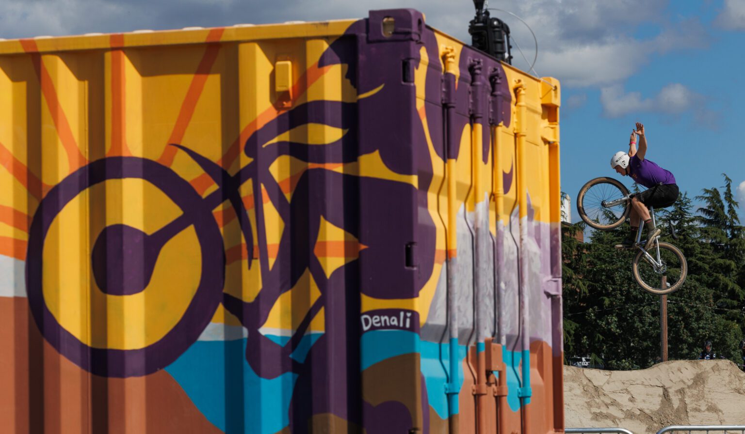 A biker poses mid-air with his bicycle near a container painted with various colors to show a biker doing tricks.