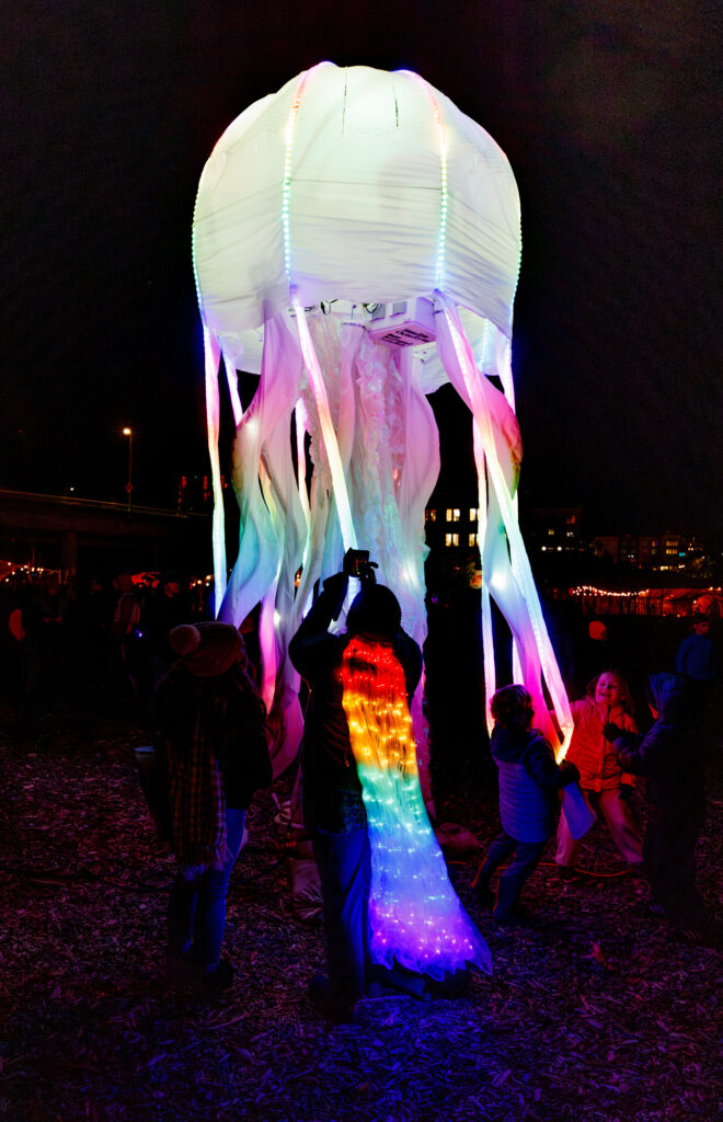 A lighted jellyfish is photographed by a person with a lighted cape.